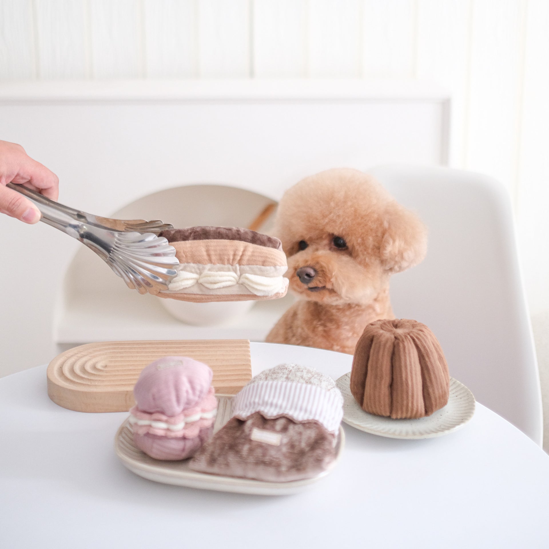 Dog Dish Lambwolf Eclair enrichment toy being held with tongs near a curious apricot poodle and pastry-themed dog toys.